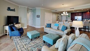 Un salon équipé d’un téléviseur, de canapés bleus et gris, de poufs, d’un tapis à motifs et d’un ventilateur de plafond.