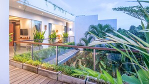Un espacio exterior moderno con terraza de madera, barandillas de vidrio y abundante vegetación.
