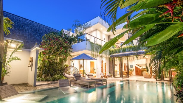 Un área de piscina moderna con sillones de descanso y un espacio de estar cubierto, rodeada de exuberante vegetación y un edificio blanco con balcones.