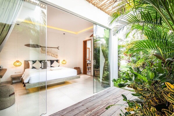 A modern bedroom with a glass wall, a wooden deck, and lush greenery outside.
