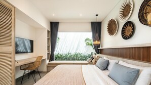 A modern bedroom with a large bed, a desk, a TV, and a view of greenery outside.