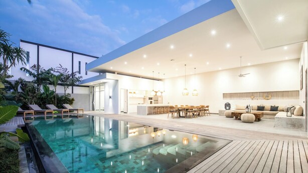 A modern outdoor pool area with lounge chairs, a wooden deck, and a well-lit living space with a sofa and dining area.