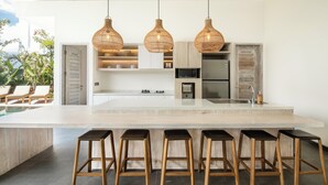 A modern kitchen with a large island, hanging wicker pendant lights, and a poolside area with lounge chairs.