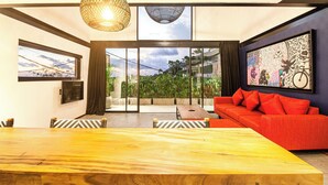 A modern living room with a wooden dining table, red sofa, and large windows offering a view of greenery.