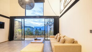 A modern living room with a sofa, ottoman, and a coffee table. A large window offers a view of a building and greenery. - Expansive Loft 200m from Canggu Beach Haven<br>Part of Noma Estate (Pererenan)
