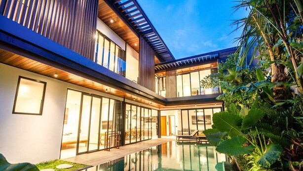 Una casa moderna con piscina cubierta por vidrio, terraza de madera y abundante vegetación.