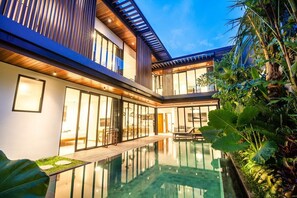 Una casa moderna con piscina cubierta por vidrio, terraza de madera y abundante vegetación.