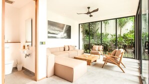 Una sala de estar moderna con un sofá seccional color beige, una mesa de centro de madera y dos sillas de madera. Hay un ventilador de techo, un ventanal con vistas a la vegetación y un cuadro en la pared.