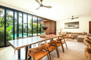 Ein geräumiges Wohnzimmer mit einem großen Holzesstisch, bequemen Sitzgelegenheiten, einer Couch und Blick auf einen Swimmingpool und üppiges Grün.
