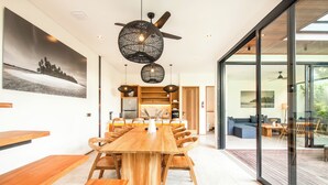 A modern dining area with a wooden table and benches, a ceiling fan, and a large window with a view of the outdoors. - Beachside Bliss: New 5BR Villa Canggu (Pererenan)