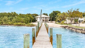 A wooden pier leading to a house with a balcony, surrounded by lush greenery and a clear blue sky. - Oceanfront Oasis: Dockside Bliss (Tavernier)
