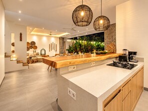 A modern kitchen with a central island, pendant lighting, and a view of a lush indoor garden. - Coastal Comfort: Cozy 4BR Villa, 300m to Finns (Kecamatan Kuta Utara)