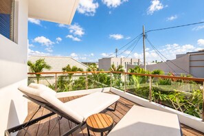 A balcony with a wooden deck, white lounge chairs, and a glass railing overlooking a residential area with palm trees and buildings.