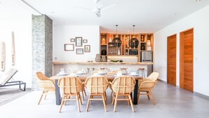 Een moderne eetruimte met een lange tafel voor zes personen, omringd door rieten stoelen. In de achtergrond is een keuken zichtbaar met houten kastjes en een inbouwoven.