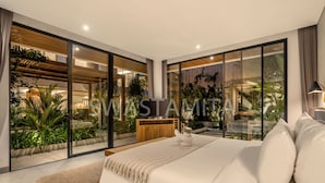 Ein modernes Schlafzimmer mit einem großen Bett, einem Holz-Kopfteil und Blick auf einen üppigen Garten durch raumhohe Glasfenster.