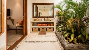 A modern bathroom with a large mirror, a sink, and a shower area. There is a wooden door leading to another room, and a small seating area with a chair and cushions. - Luxurious 5BR Dream Villa, 200m to Berawa Beach (Canggu)