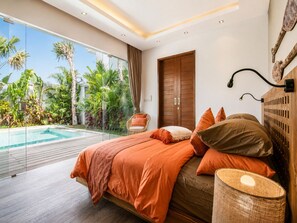 Un dormitorio con una cama grande, cabecera de madera, lámpara de noche y vistas a una piscina y plantas tropicales.