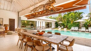 A modern indoor pool area with a wooden dining table and chairs, a unique hanging light fixture, and a view of the outdoor pool with lounge chairs. - Luxurious 5BR Dream Villa, 200m to Berawa Beach (Canggu)