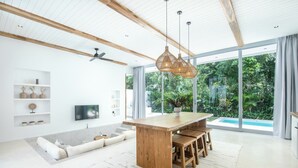 A modern living room with a wooden dining table, built-in shelves, a flat-screen TV, and a large sliding glass door leading to a pool. - New 2br villa 4mins to Bingin beach (Uluwatu)