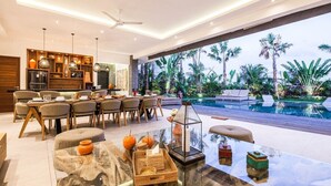 A modern living room with a glass coffee table, a dining area with wooden chairs, and a view of a pool with palm trees. - Splendid 6BR Villa, Just 400m to the Shoreline (Kuta Utara)