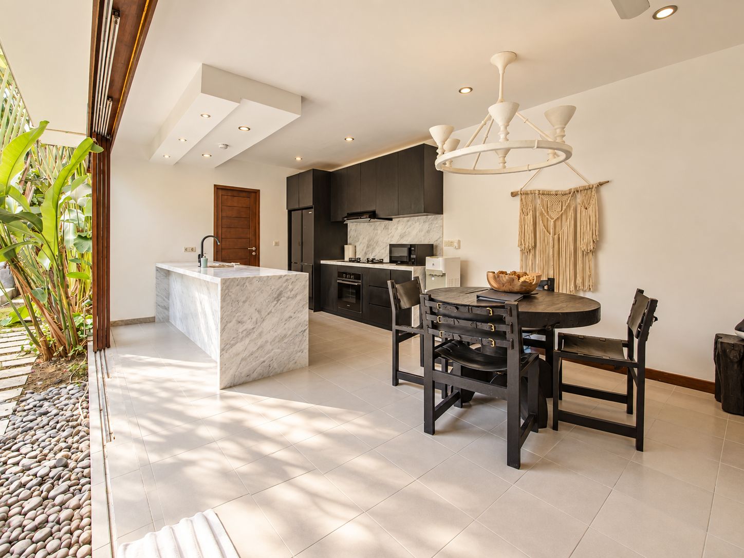 A modern kitchen with a marble island, dark cabinetry, and a dining area with a round table and chairs.