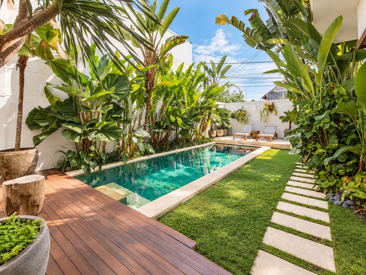 Une piscine entourée d’une végétation luxuriante, une terrasse en bois et un chemin pavé de pierres.