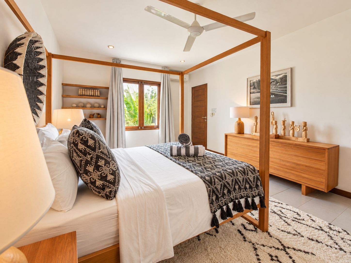 A bedroom with a canopy bed, a wooden dresser, a desk with a lamp, and a window with curtains.