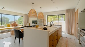 Una cocina moderna con una isla grande, luces colgantes y vistas al exterior con vegetación.