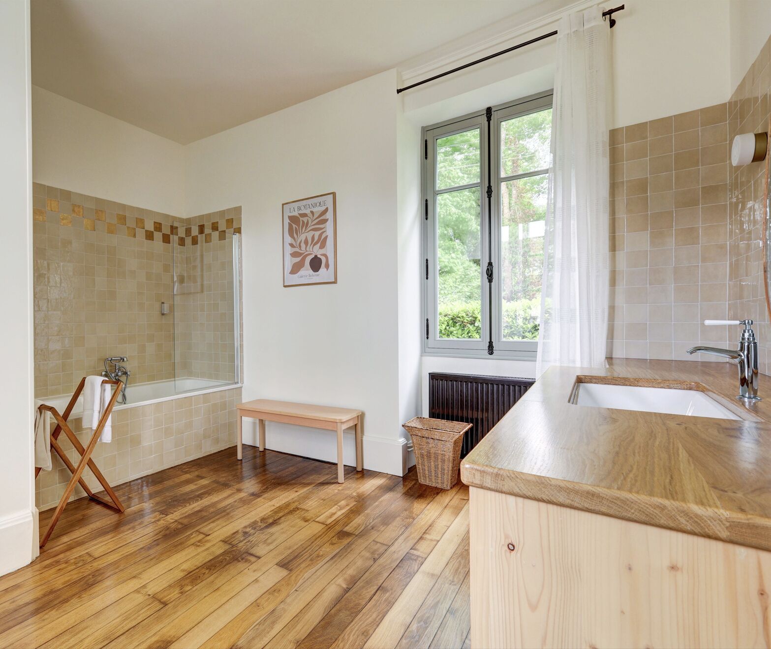 Un baño con una bañera, un banco de madera, una cesta de mimbre y una ventana con vistas a la vegetación.