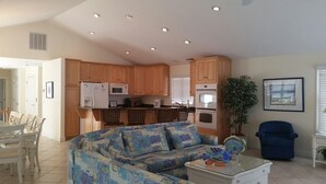 A modern kitchen with wooden cabinets, a dining area with a white table and chairs, and a living room with a blue armchair and a sofa. - Bethany Beach 6 BR with Elevator - one house back from the beach (Bethany Beach)
