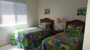 Un dormitorio con dos camas individuales, una ventana con persianas, una mesita de noche con lámpara y dos cuadros en la pared.