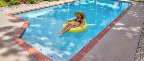 A swimming pool with a person on a yellow float, surrounded by a brick border and greenery.