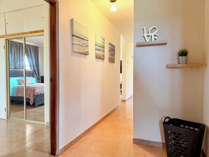 A hotel corridor with a bed visible through a glass door, artwork on the walls, a shelf with a plant, and a trash bin.