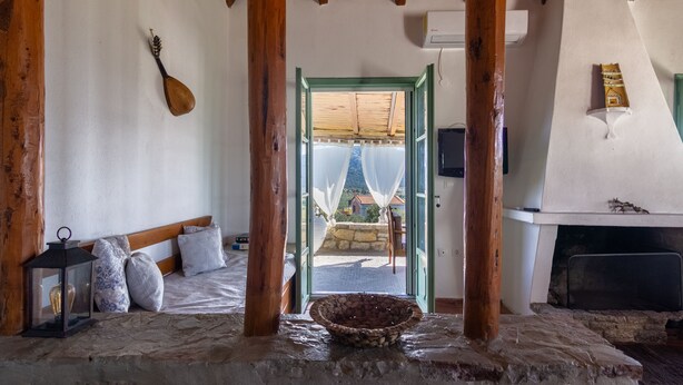 Una habitación rústica con piso de piedra, vigas de madera, una cama con almohadones, un farol, un lavabo de piedra y vistas a un balcón desde un umbral abierto.