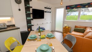 A modern kitchen with a flat-screen TV, a dining table set with teal plates, and a yellow chair. - Nanny's Little House (Sandown)