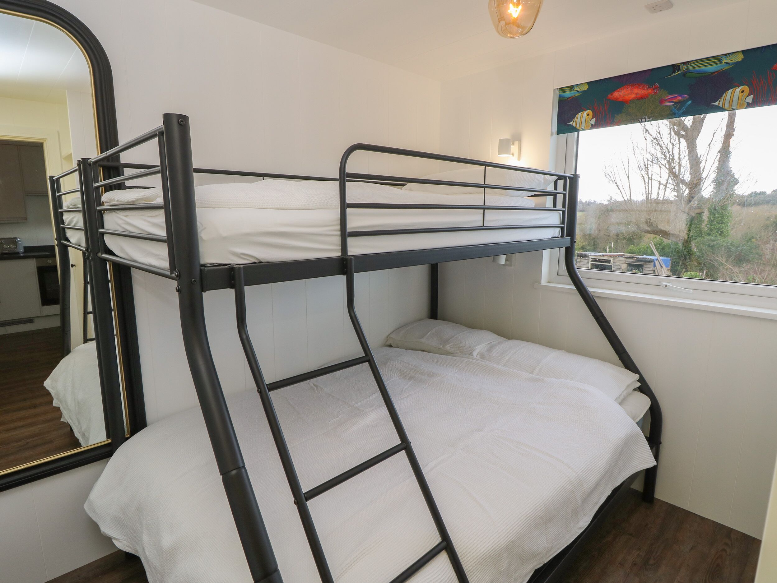 A bunk bed with a ladder, white bedding, a window with a view of trees, and a wall-mounted light fixture.