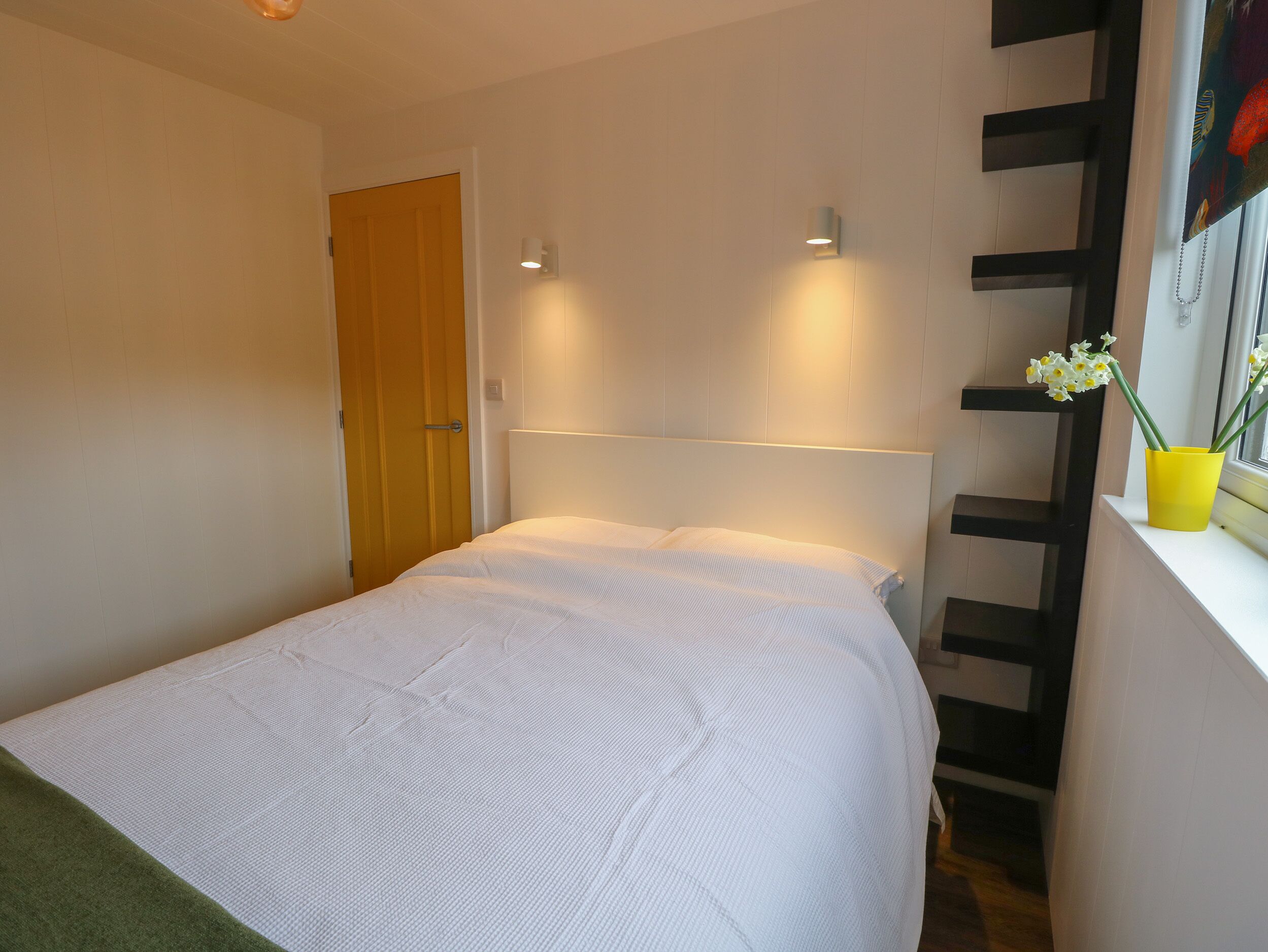 Een eenpersoonsbed met een wit matras en hoofdeinde, een houten deur en een raam met een gele bloevas.