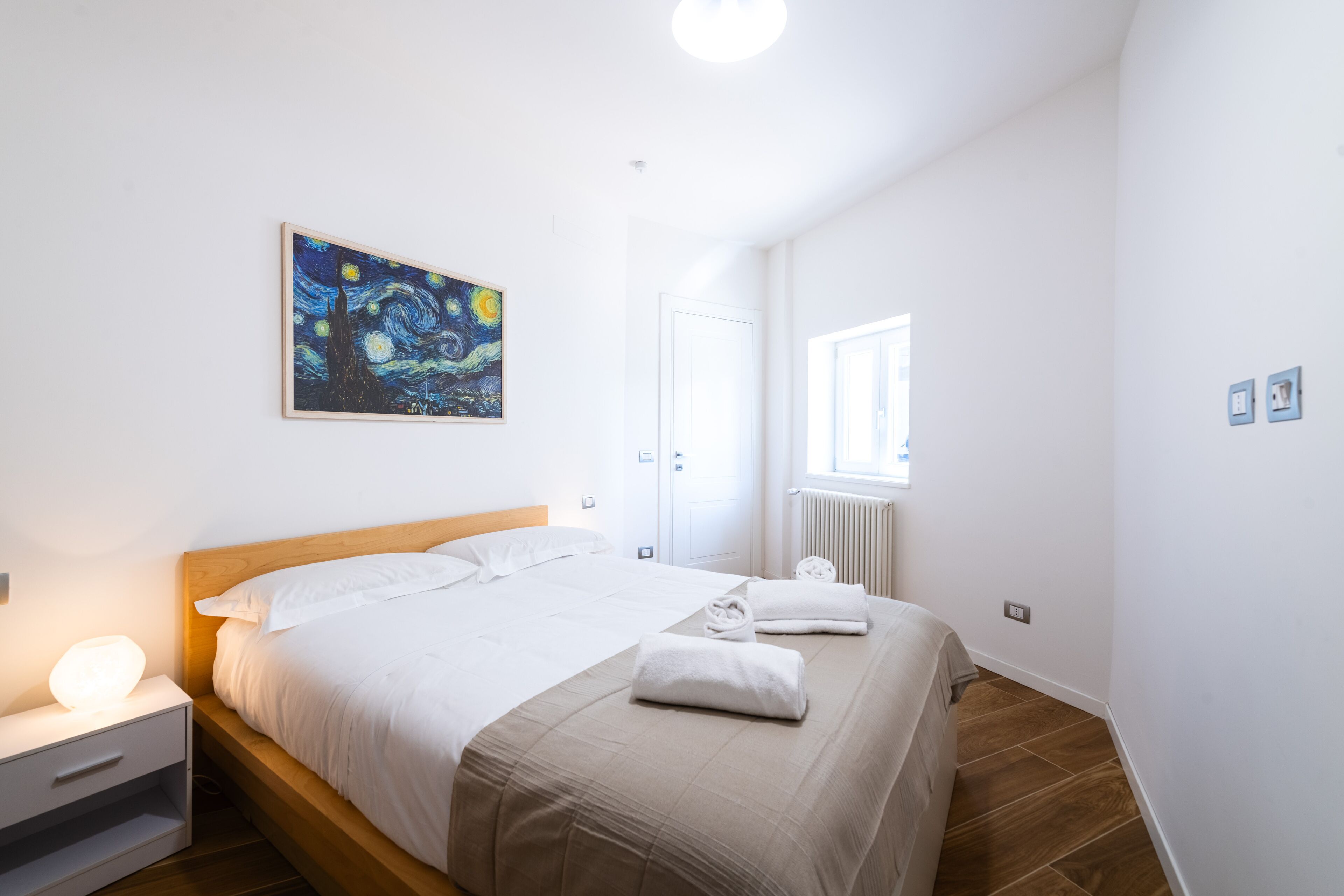 Un dormitorio con cama, mesita de noche, cuadro, ventana y radiador.