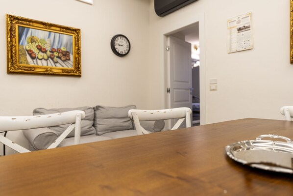 A dining area with a wooden table, white chairs, a painting, a clock, and a door leading to another room.