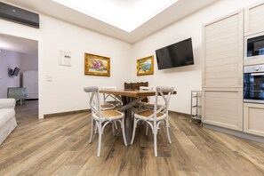 A dining area with a wooden table and white chairs, a TV mounted on the wall, and a kitchen area with a built-in oven. - Plebiscito Rooms - Camera 2 (Naples)