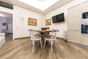 Un coin repas avec une table en bois et des chaises blanches, un téléviseur fixé au mur et un espace cuisine doté d’un four encastré.