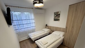 A small, clean room with two single beds, a bedside table with a lamp, a wall-mounted TV, and a window with sheer curtains. - Május 1 Vendégház - Torony (Torony)