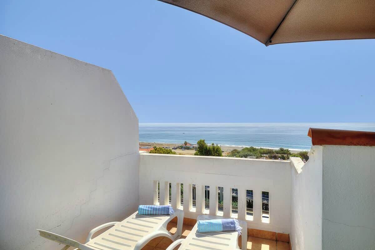 A balcony with two white chairs and a beige umbrella, overlooking a beach and ocean.