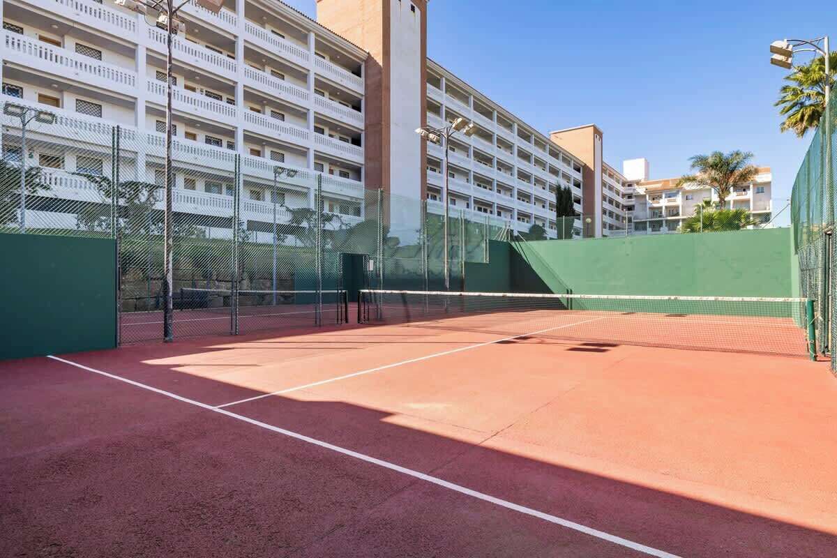 Uma quadra de tênis com rede, cercada por uma grade, e adjacente a um edifício com varandas.