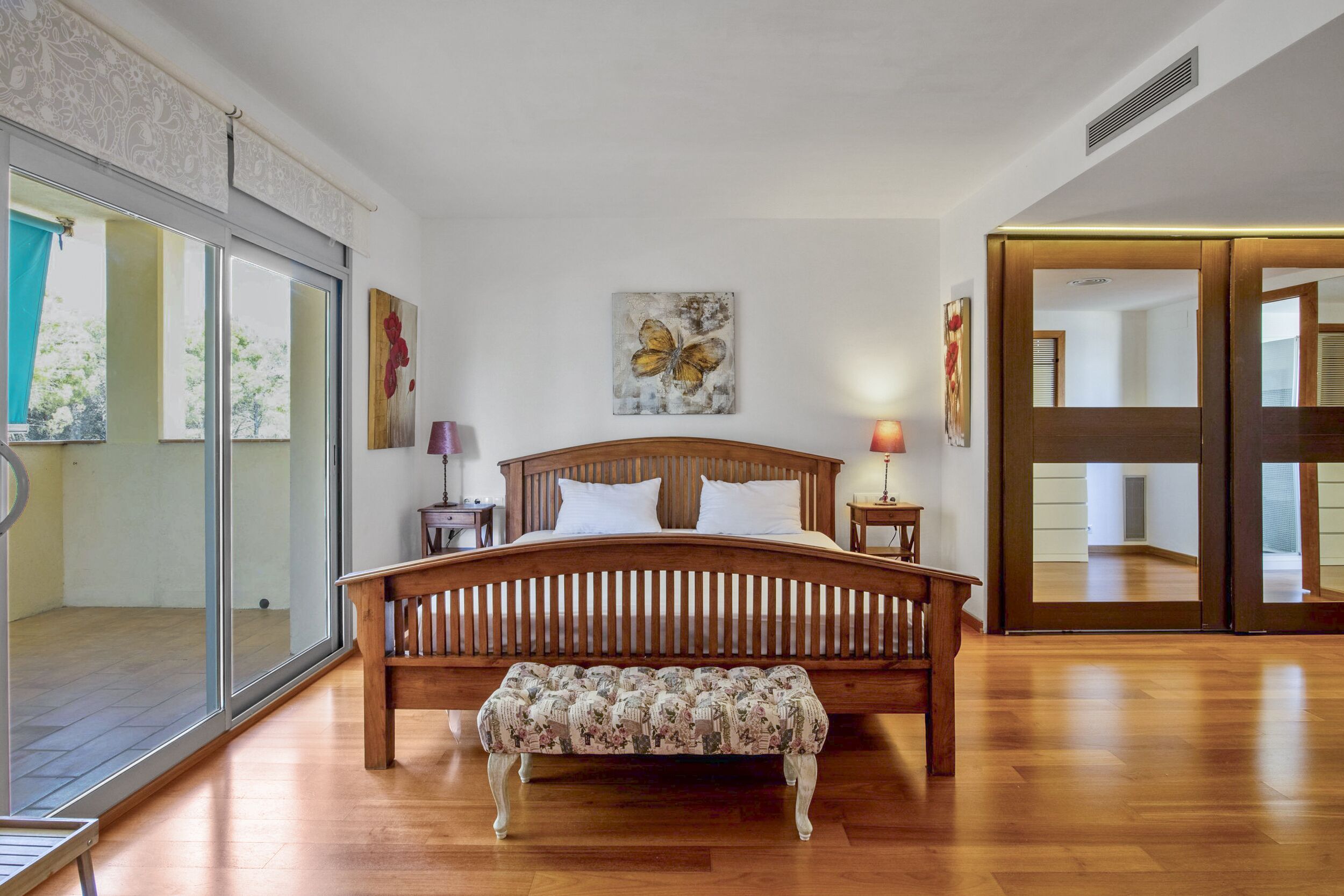 A bedroom with a wooden bed, a bench, a window with a view, and artwork on the walls.