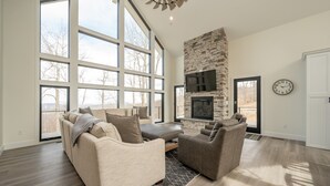 A spacious living room with large windows, a stone fireplace, and a flat-screen TV.