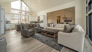 A spacious living area with a large sofa, two armchairs, and a coffee table. There is a kitchen in the background with white cabinets and a large window.