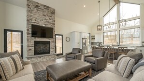 A spacious living room with a stone fireplace, comfortable seating, and large windows offering a view of the outdoors.