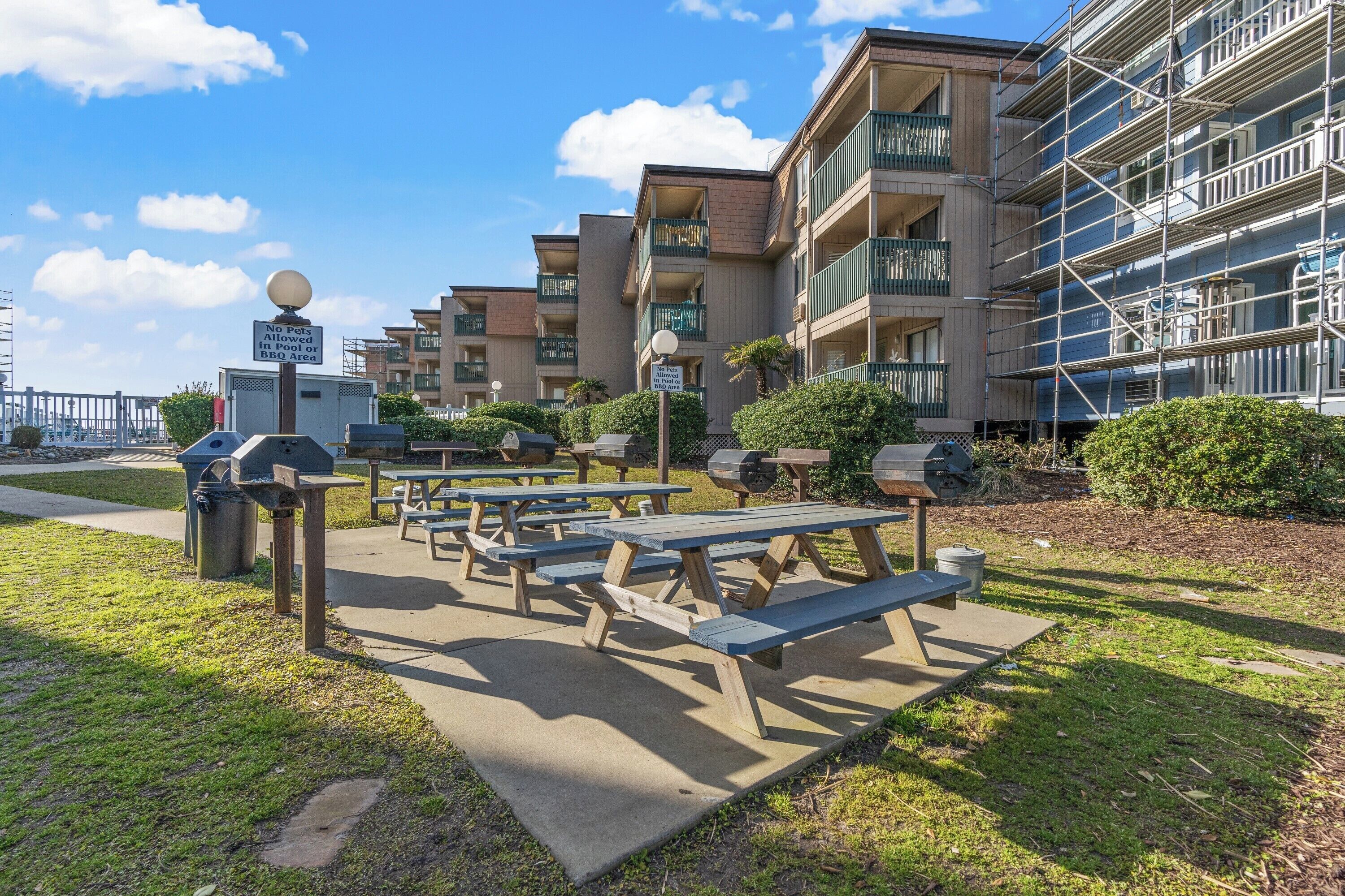 Outdoor picnic tables, a BBQ grill, and a no pets allowed sign in front of a multi-story building with balconies.