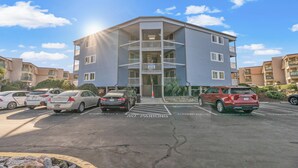 A multi-story residential building with a central entrance, parking lot, and a clear sky. - Oceanfront View @ A Place at the Beach 3 1I (Myrtle Beach)